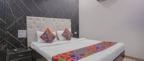Un dormitorio con tablero de cabecera capitonado, cama con ropa de cama blanca y almohadas geométricas de colores, mesita de noche con control remoto y aire acondicionado instalado en la pared.