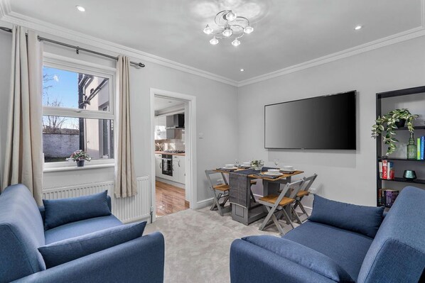 A modern living room with a dining area, a flat-screen TV mounted on the wall, and a bookshelf with books and decorative items.