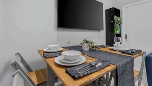 A dining area with a wooden table set for two, white plates, cutlery, and glasses. A flat-screen TV is mounted on the wall above the table. - East Croydon Home, Near Station & Gatwick Airport (Greater London)