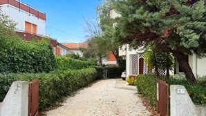 Uma estrada de cascalho leva a uma casa com portão, cercada por vegetação e um céu claro.