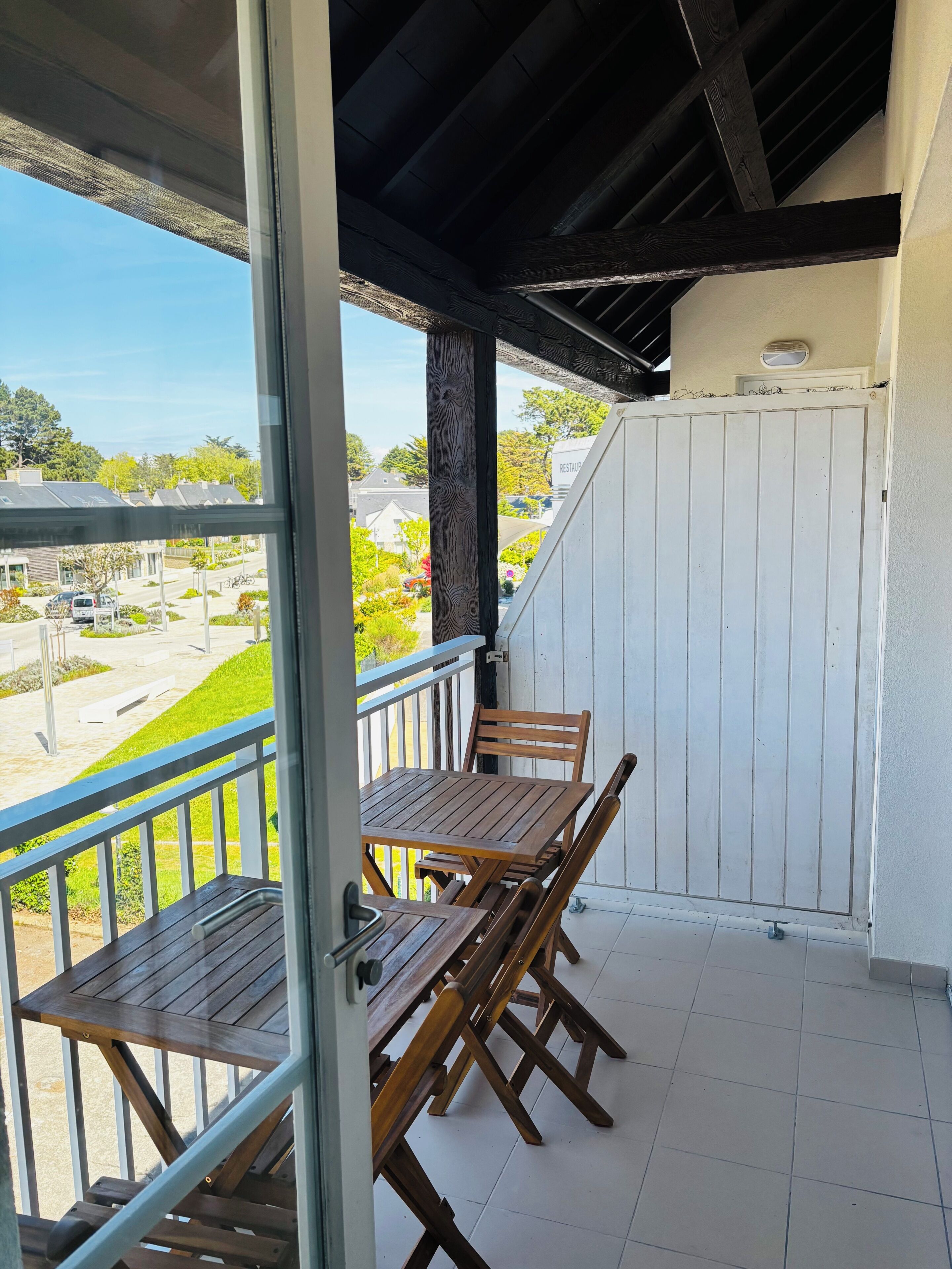 Un balcón con mesa y sillas de madera, un portón blanco y vistas a una zona residencial.