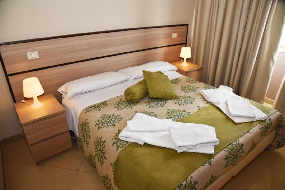 Cama bem arrumada com almofadas, abajur e toalha dobrada sobre a cama.