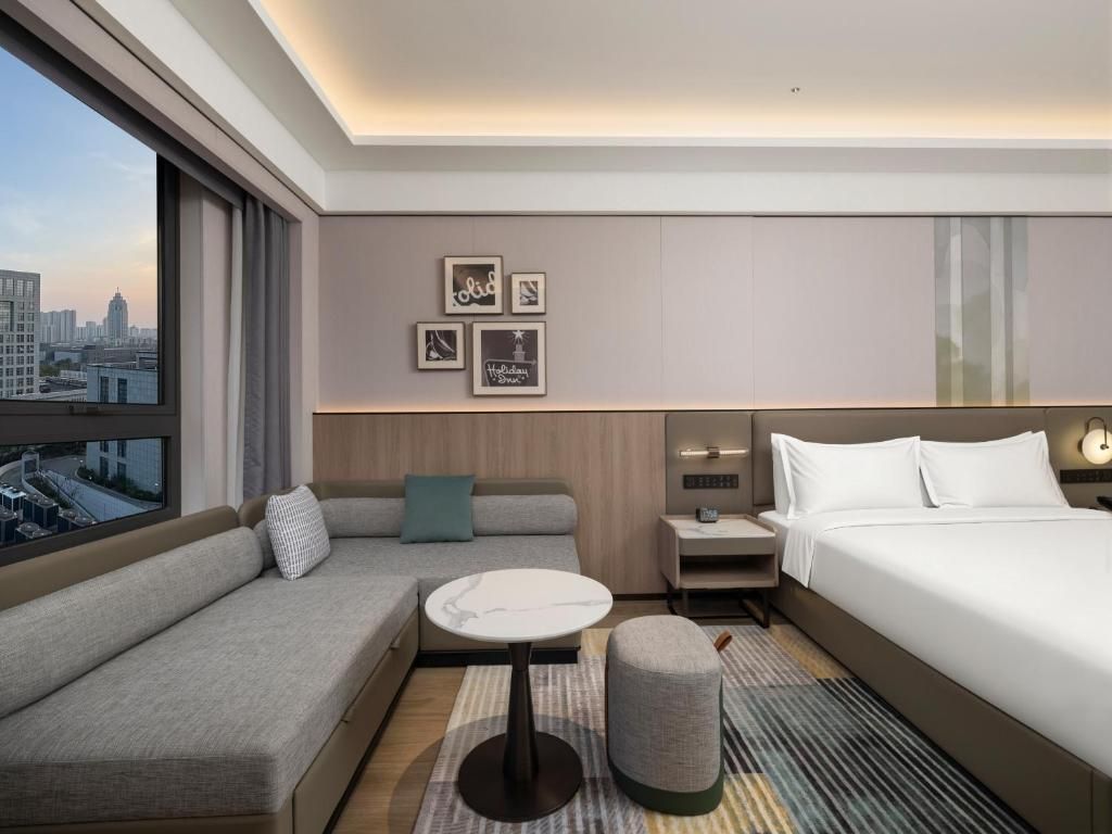 A modern hotel room with a sofa, a small table, a bed, a nightstand, and a city view.