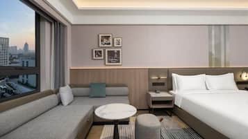 A modern hotel room with a sofa, a small table, a bed, a nightstand, and a city view.