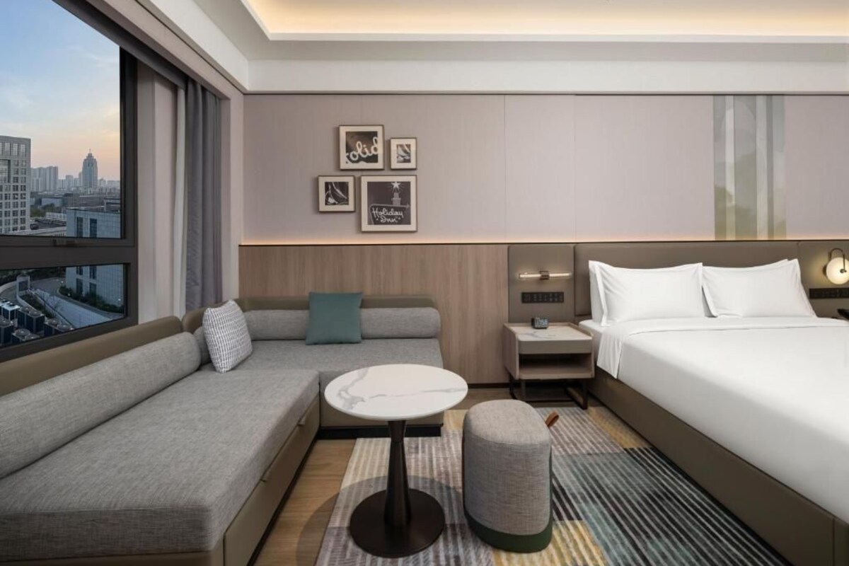 A modern hotel room with a sofa, a small table, a bed, a nightstand, and a city view.
