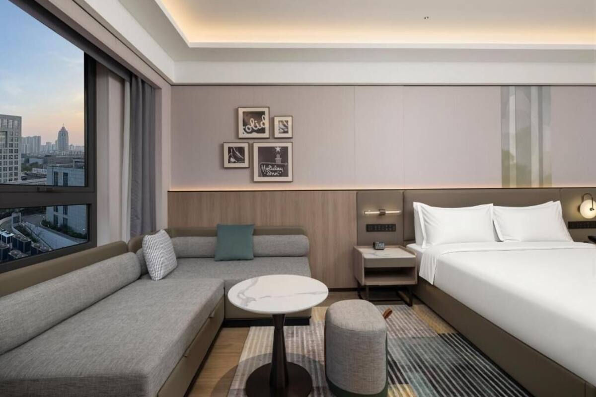 A modern hotel room with a sofa, a small table, a bed, a nightstand, and a city view.