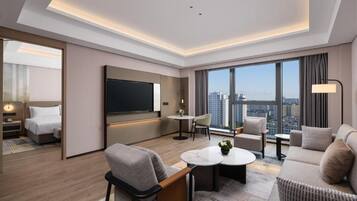 Habitación de hotel moderna con vistas a la ciudad, televisor de pantalla plana, sofá, mesa de centro y un pequeño comedor.