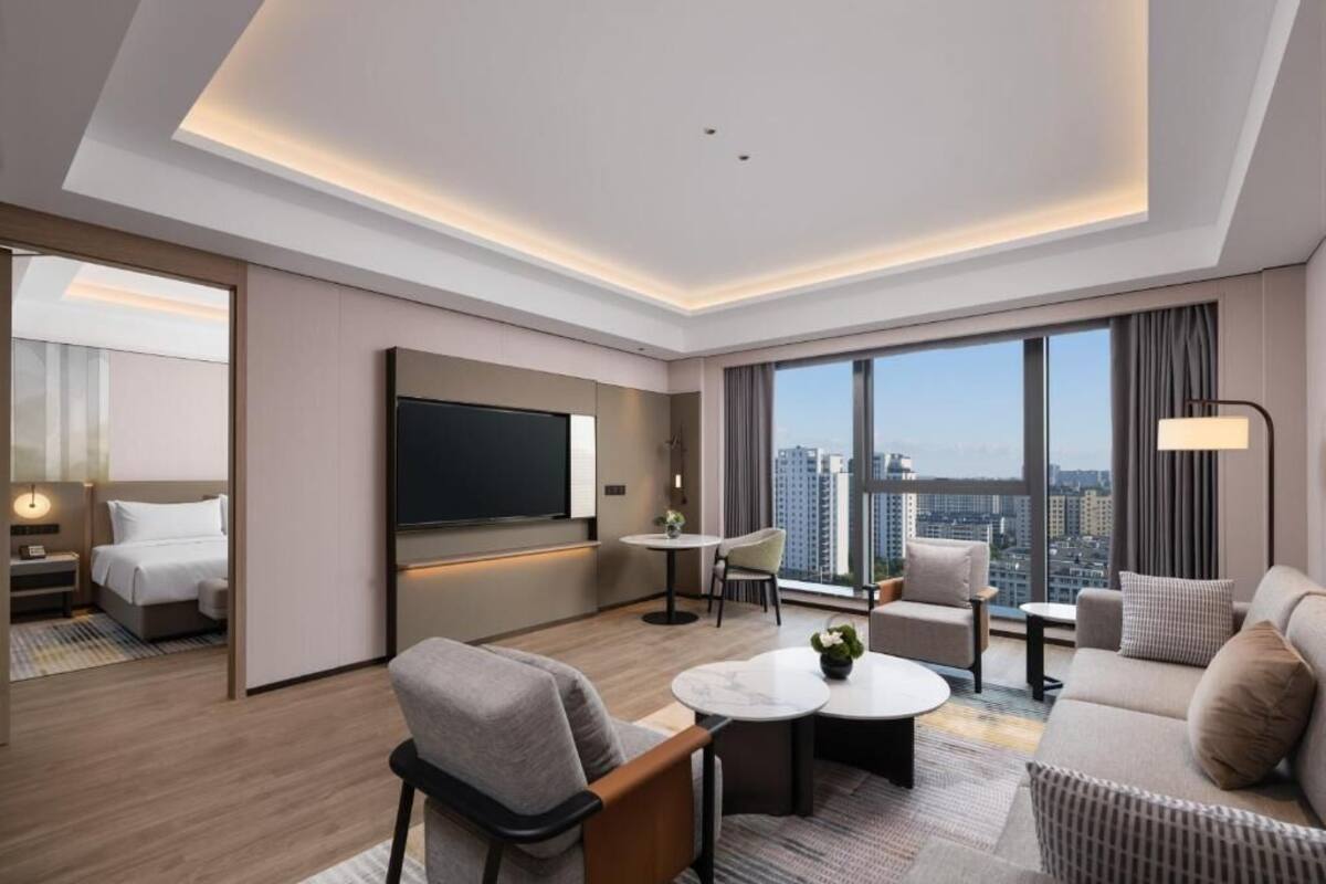 A modern hotel room with a city view, a flat-screen TV, a sofa, a coffee table, and a small dining area.