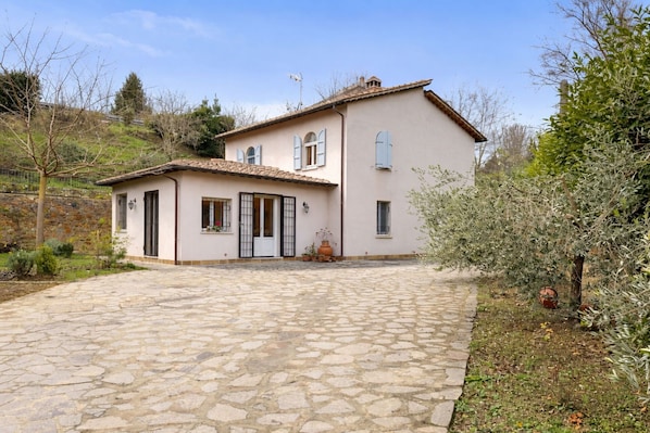 A two-story house with a stone-paved driveway, a small garden, and a stone wall in the background.