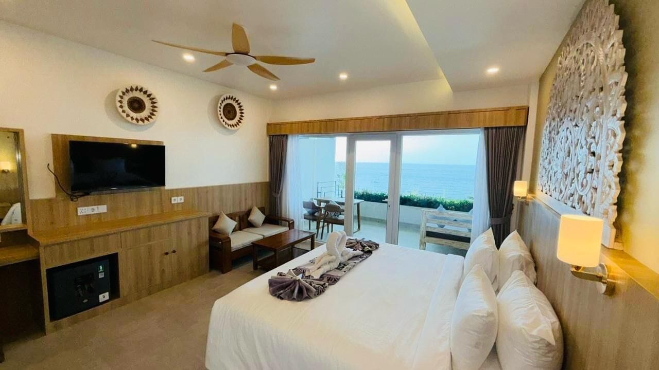 A hotel room with a large bed, a TV, a ceiling fan, a view of the sea, and a balcony with outdoor furniture.