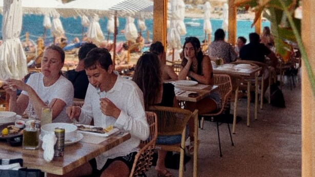 An outdoor dining area with tables and chairs, umbrellas, and a view of the sea and hills in the background. - Hotel Porta Ksamil (Ksamil)