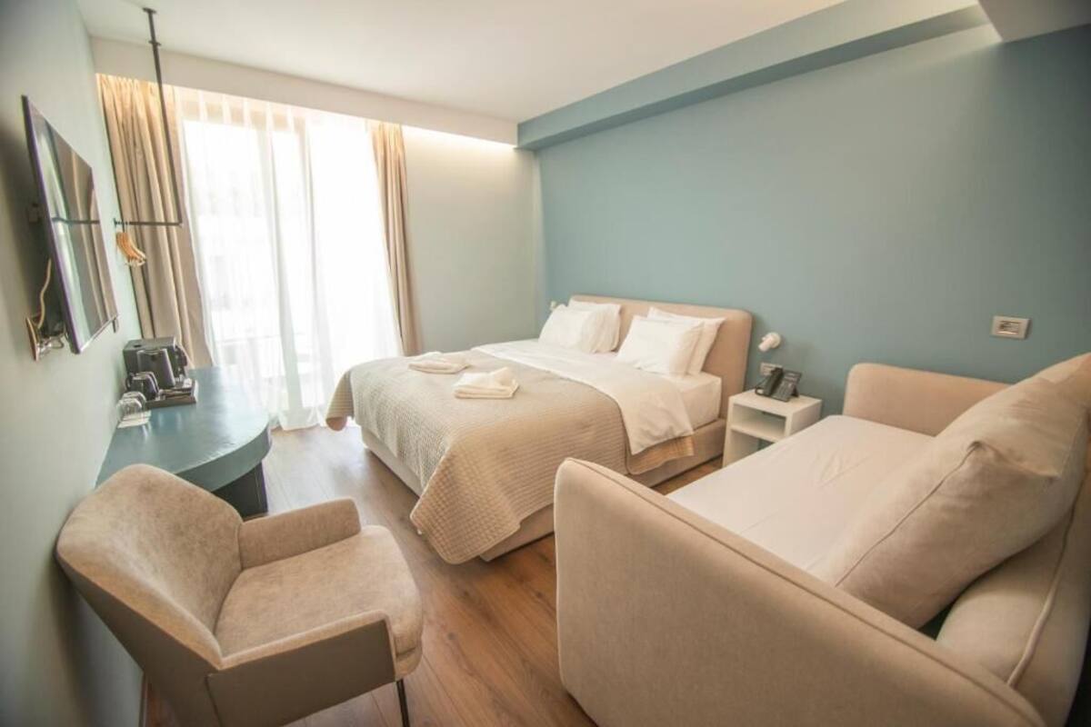 Habitación de hotel con cama, sofá, sillón, escritorio con lámpara y ventana con cortinas.