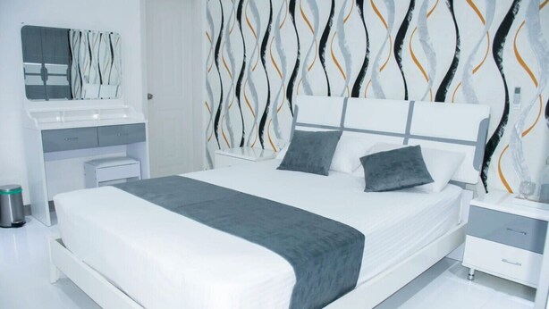 Un dormitorio moderno con una cama blanca, almohadas grises, una manta gris, una mesita de noche blanca y un buró.