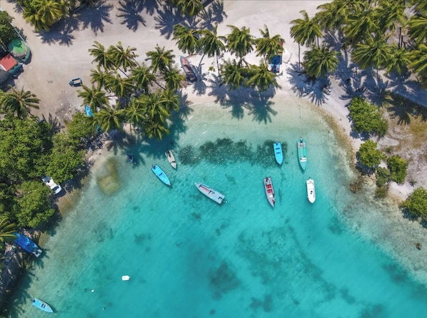 An aerial view of a beach with several boats anchored near the shore, surrounded by palm trees and clear turquoise water.