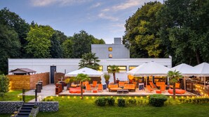 A modern outdoor lounge area with seating and umbrellas, a wooden fence, and a staircase leading to a building.