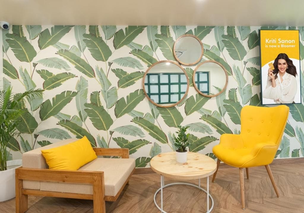 A room with a tropical leaf-pattern wallpaper, a wooden sofa with a yellow cushion, a round wooden table with a potted plant, and a bright yellow chair.