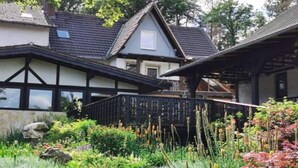 Une maison traditionnelle avec un toit à pignon, une terrasse en bois et un jardin agrémenté de diverses plantes et fleurs.