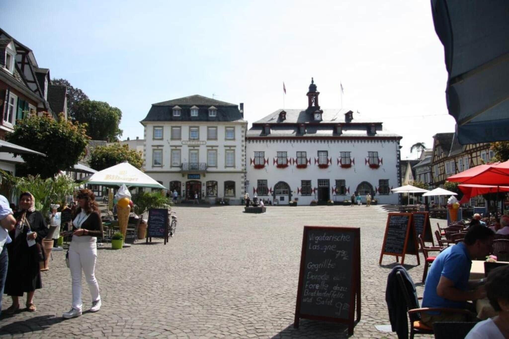 Ein Café mit Außenbereich, Tischen und Stühlen, einem gepflasterten Boden und einem Gebäude mit weißer Fassade und roten Akzenten im Hintergrund.