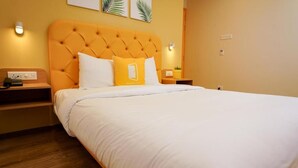 Una habitación de hotel con una cama grande, un cabecero naranja con capitonado y una colcha blanca. Hay dos mesitas de noche, con lámparas y una pequeña planta en una de ellas.