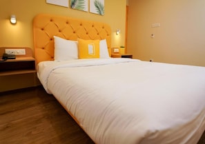 Una habitación de hotel con una cama grande, un cabecero naranja con capitonado y una colcha blanca. Hay dos mesitas de noche, con lámparas y una pequeña planta en una de ellas.