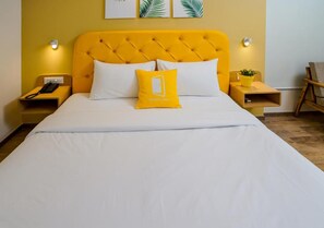 A bed with a yellow headboard, white bedding, and a yellow pillow with a logo. There are two bedside tables with lamps and a small plant.