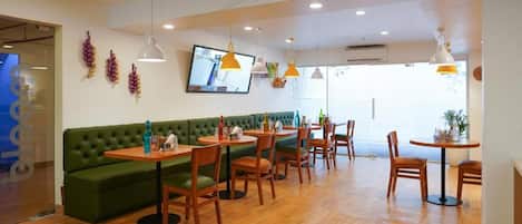 A restaurant with wooden tables and chairs, a green sofa, pendant lights, and a television on the wall.