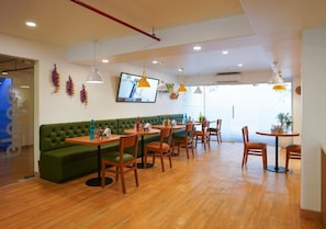 A restaurant with wooden tables and chairs, a green sofa, pendant lights, and a television on the wall. - Bloom Hotel - Koregaon Park (PUNE)