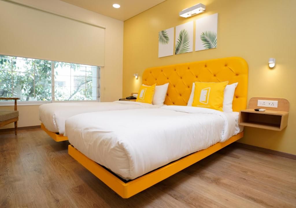 Cama doble con cabecera amarilla, ropa de cama blanca y dos almohadas amarillas. Hay una mesita de noche con una lámpara, un enchufe y un pequeño estante. La habitación tiene suelo de madera, un ventanal con vistas a árboles y una lámpara fijada a la pared.