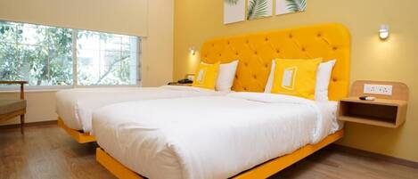 A double bed with a yellow headboard, white bedding, and two yellow pillows. There is a bedside table with a lamp, a power outlet, and a small shelf. The room has wooden flooring, a large window with a view of trees, and a wall-mounted light fixture.