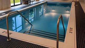 An indoor swimming pool with clear water, a tiled deck, and a seating area with chairs and tables.