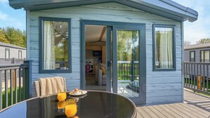 Een modern, compact vakantiehuisje met een glazen deur naar een keuken en eetgedeelte, met een houten terras en buiten zitplaatsen.