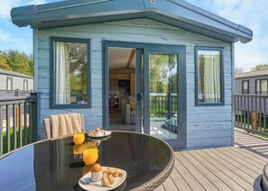 A modern, compact cabin with a glass door leading to a kitchen and dining area, featuring a wooden deck and outdoor seating. - Luxury 2 Bedroom Hot Tub sleeps 6 (Hayle)