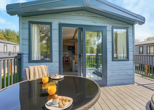 Una cabaña moderna y compacta con puerta de vidrio que da a una cocina y zona de comedor, con terraza de madera y asientos exteriores.