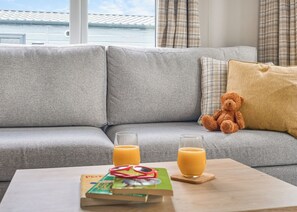 A grey sofa with a teddy bear, a glass of orange juice, and a book on a wooden table. - Luxury 2 Bedroom Hot Tub sleeps 6 (Hayle)