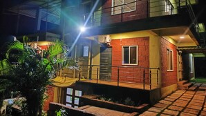 A two-story building with a balcony on the upper level, a small porch area with a chair, and a window on the ground floor. - Red Soil Strawberry Farm villa (Bhuteghar)