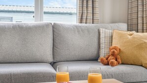 Um sofá cinzento com um urso de pelúcia, um copo de sumo de laranja e um livro sobre uma mesa de madeira.