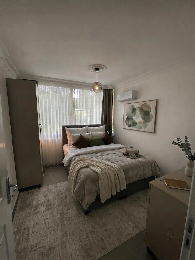 Een slaapkamer met een bed, nachtkastje, raam met gordijnen en een wandairconditioner.