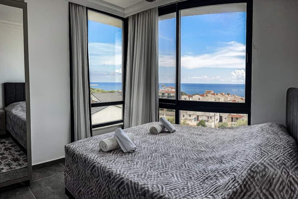 Ett hotellrum med en säng, ett fönster med utsikt över byggnader och havet, och en stol.