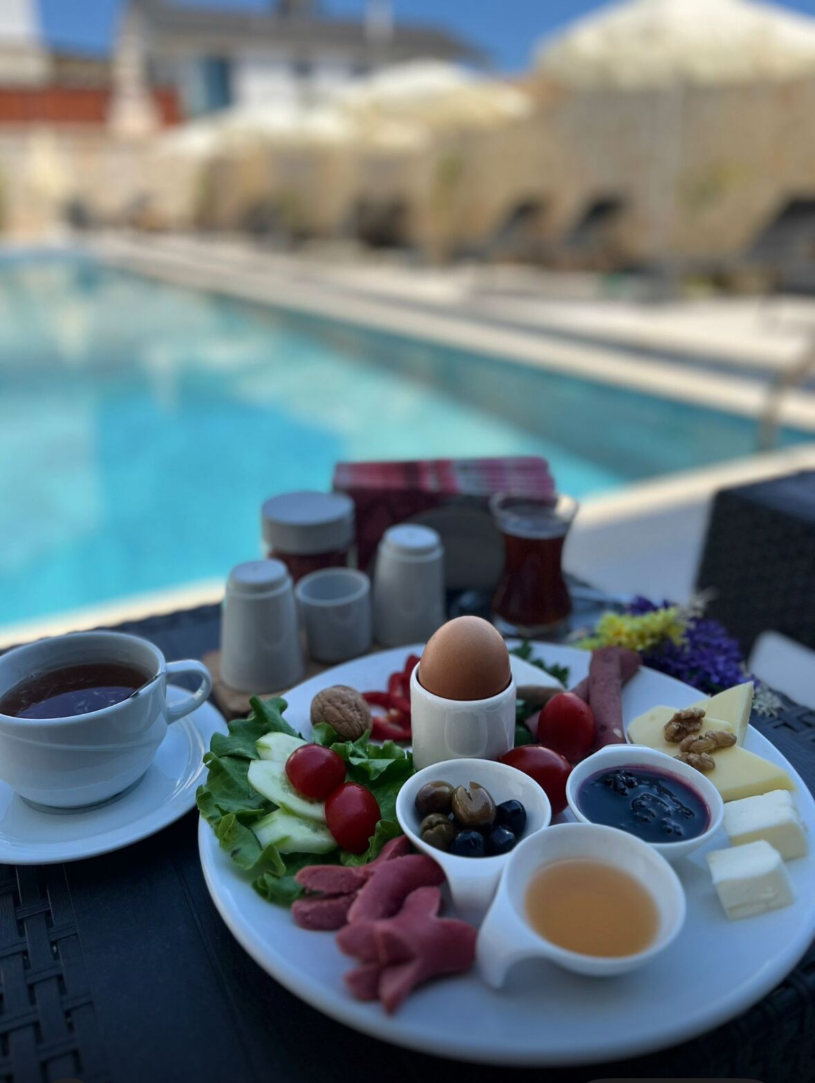 A table with a plate of food, a cup of coffee, and a pool in the background.