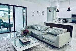 Una sala de estar moderna con un sofá seccional gris, una mesa de centro de vidrio con un florero, y una zona de cocina con luces colgantes y un microondas.