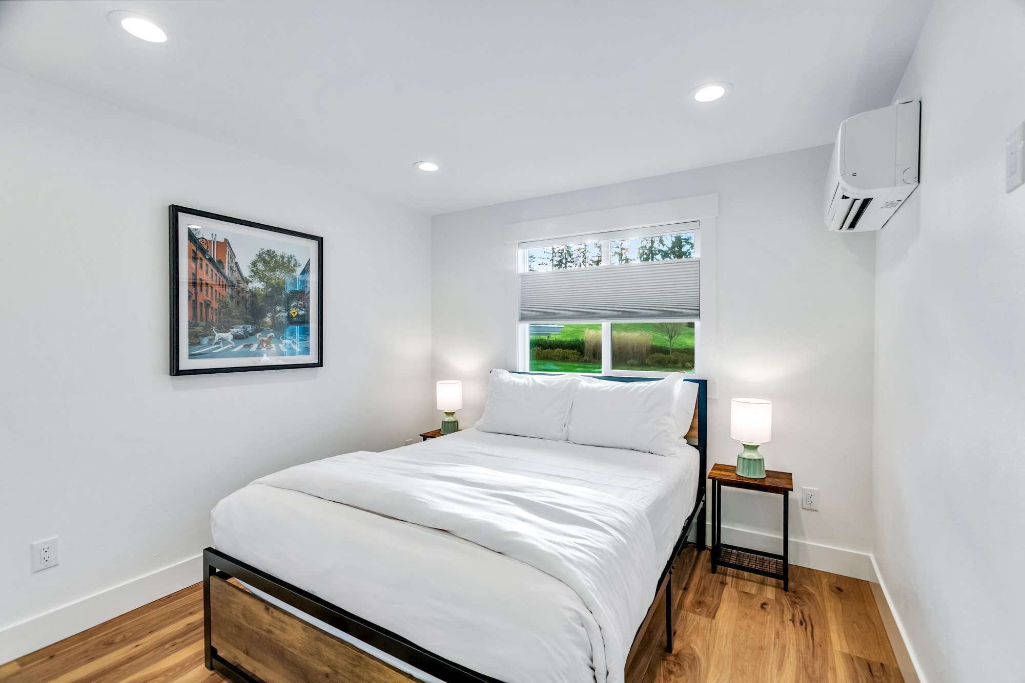 A modern bedroom with a bed, two bedside tables with lamps, a window with blinds, an air conditioning unit, and a framed picture on the wall.