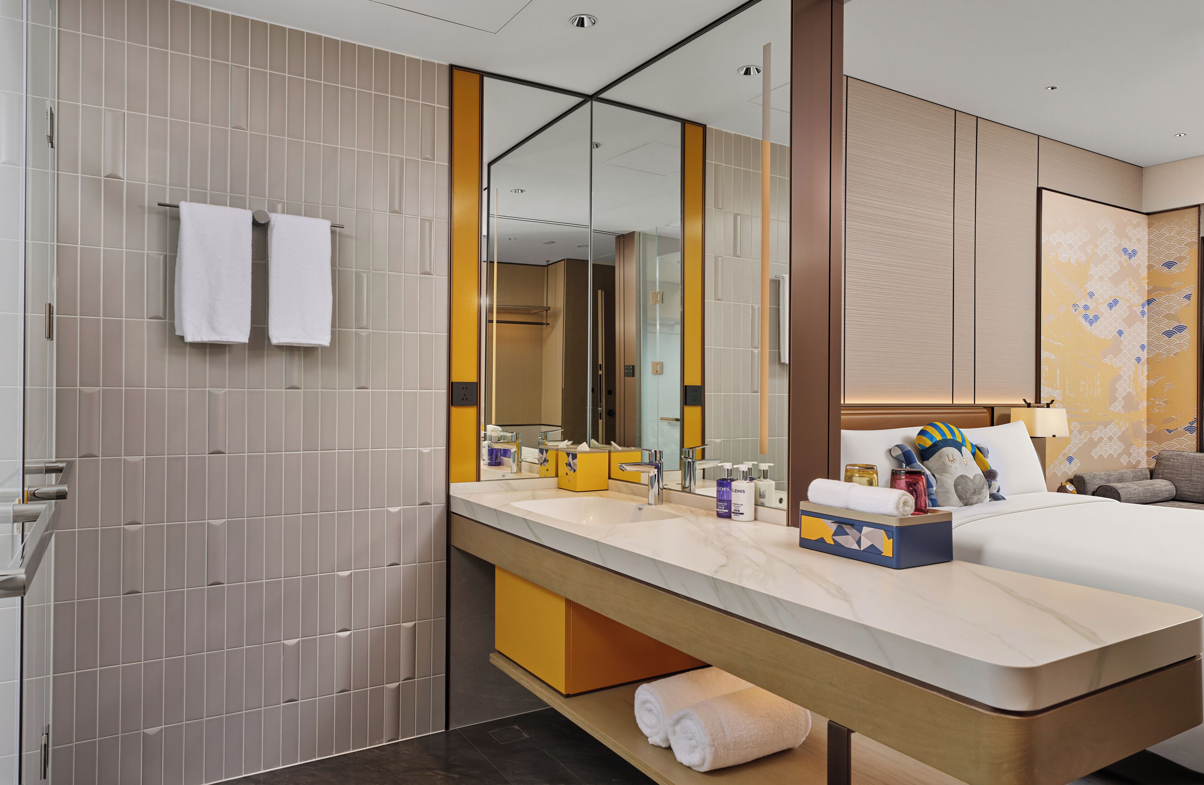Une chambre d’hôtel moderne avec un grand miroir, un double lavabo, un lit avec des oreillers et une table de chevet.
