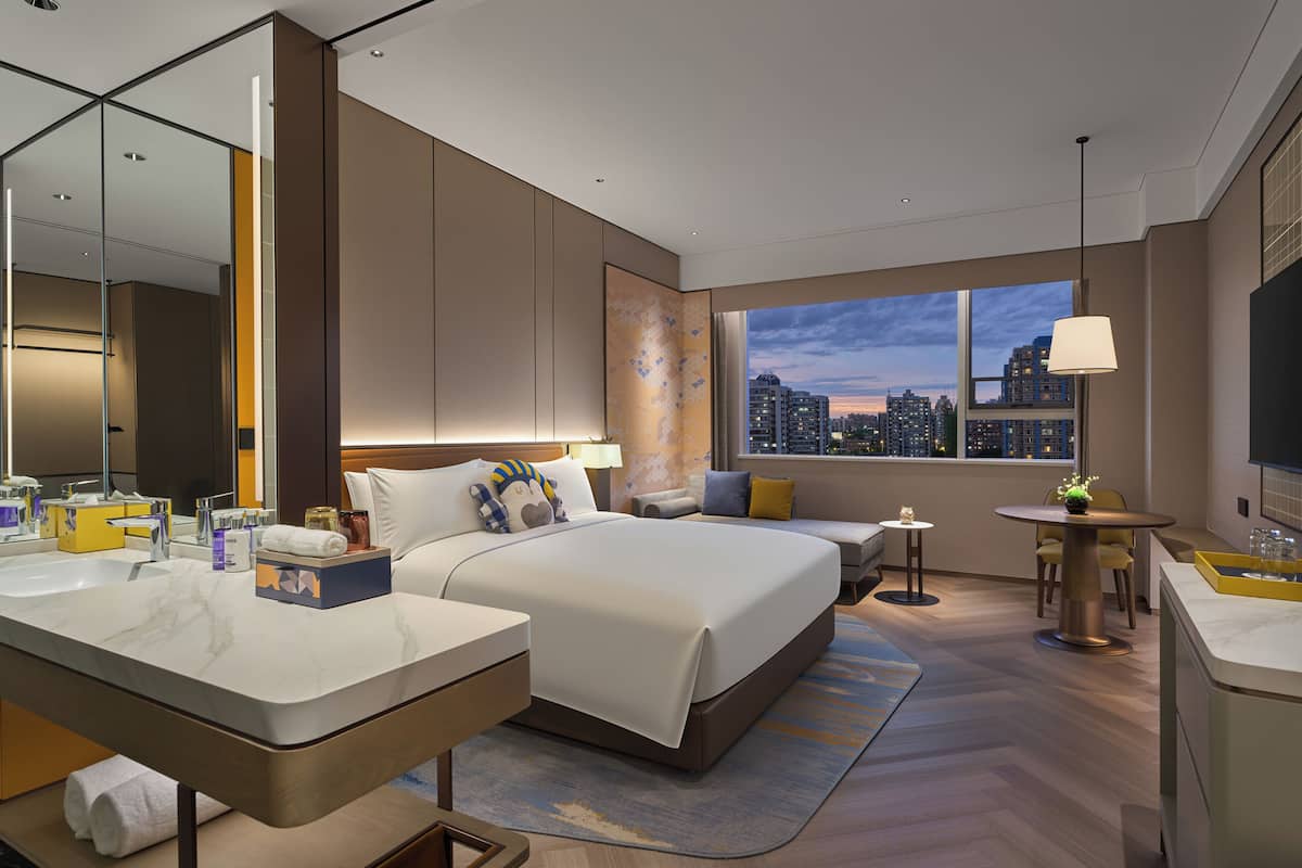 A modern hotel room with a large bed, a sitting area, a small table, a bathroom with a mirror, and a city view.