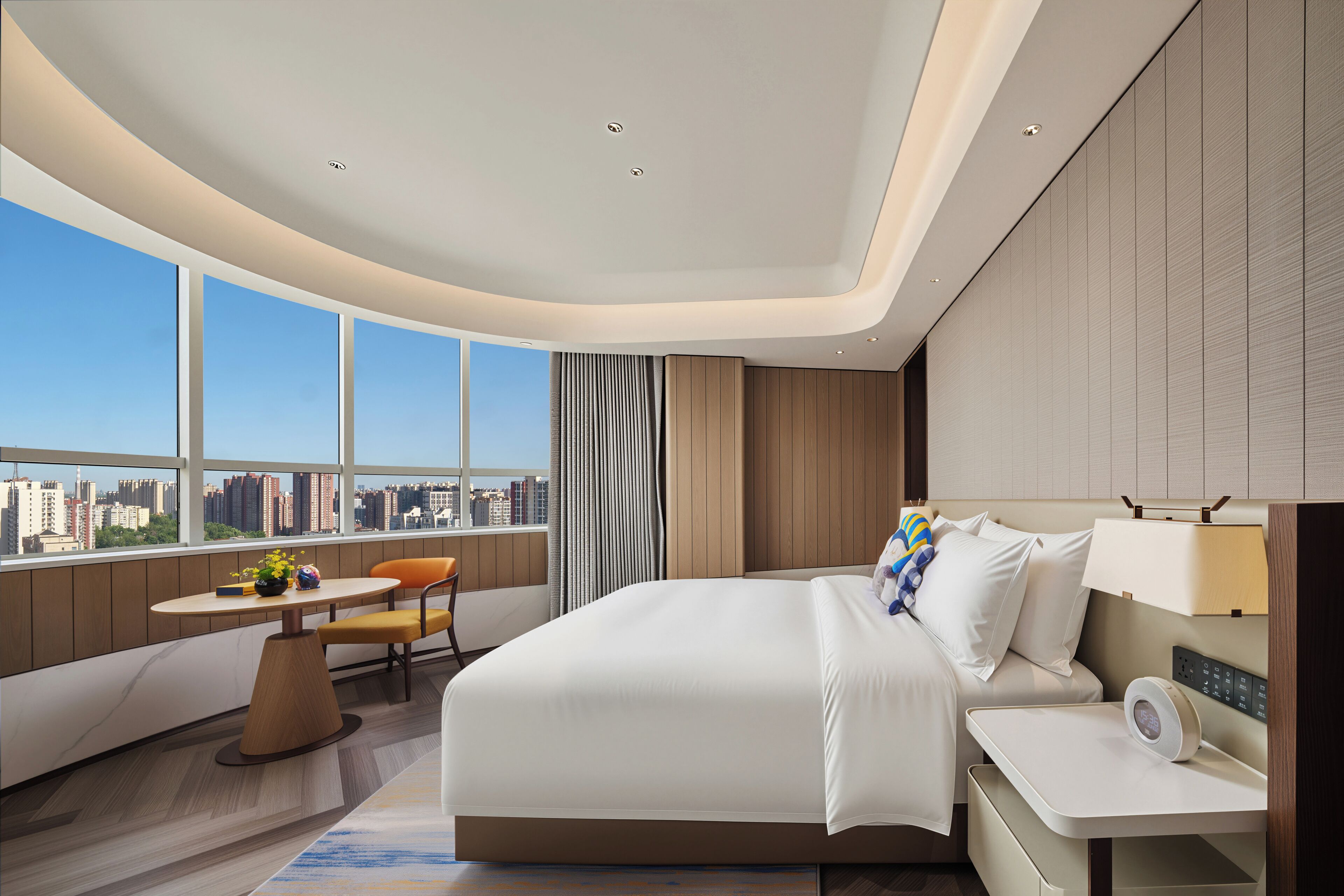 A modern hotel room with a curved ceiling, a large bed, a bedside table with a lamp, a small table with a chair, and a view of the city through large windows.