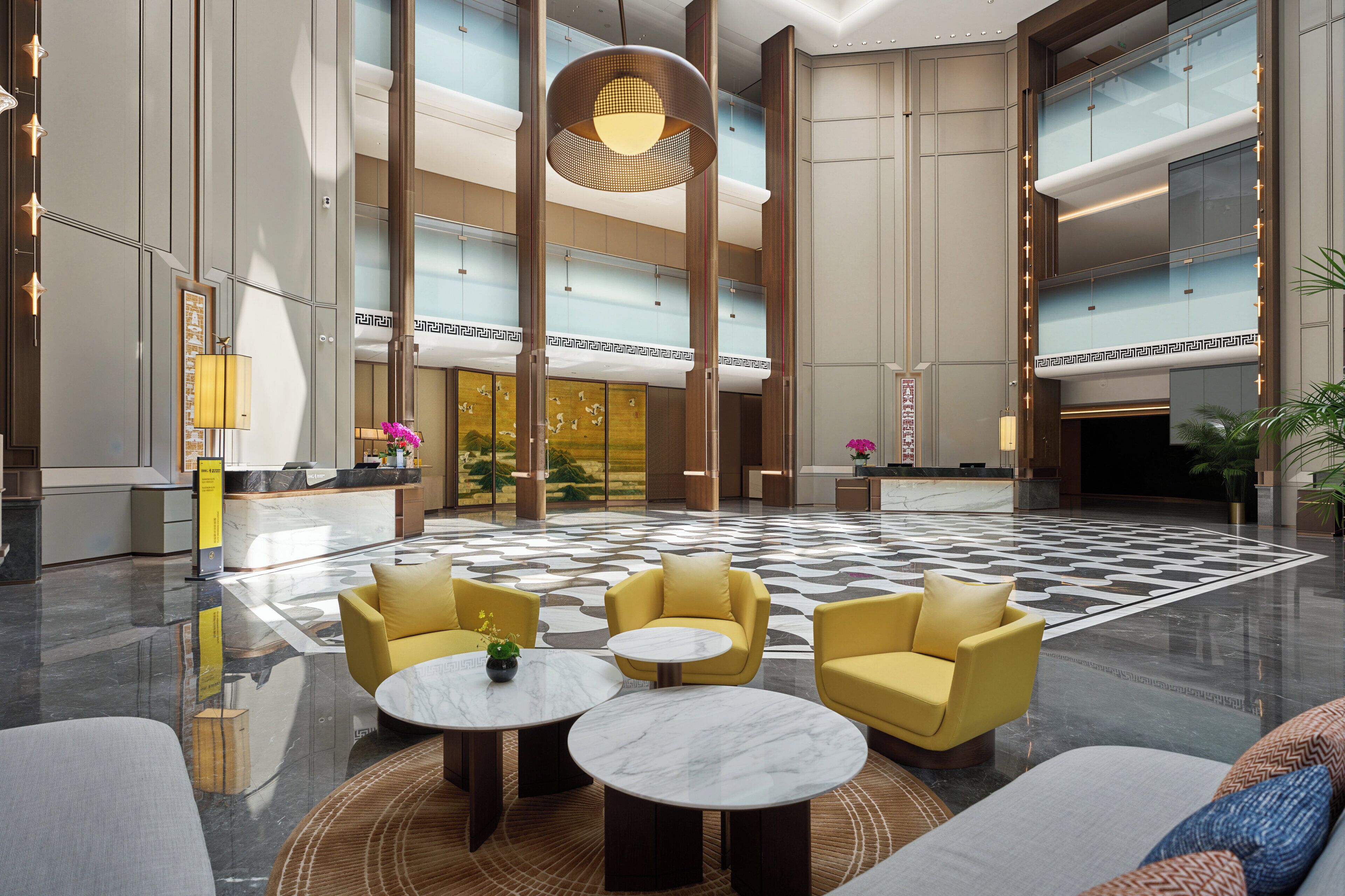 A spacious lobby with high ceilings, a checkered floor, and modern furnishings including yellow armchairs and round tables.
