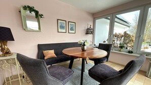 Ein gemütlicher Essbereich mit einem runden Holztisch, zwei grau gepolsterten Stühlen, einer schwarzen Couch und einem kleinen Beistelltisch mit Lampe.