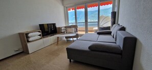 Ein modernes Wohnzimmer mit grauem Sofa, einem Flachbildfernseher, einem kleinen weißen Tisch und einem Balkon mit rotem Markisenvorzelt.