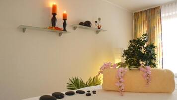Une salle de spa avec une table de massage blanche, des bougies, une plante et une étagère ornée d’objets décoratifs.