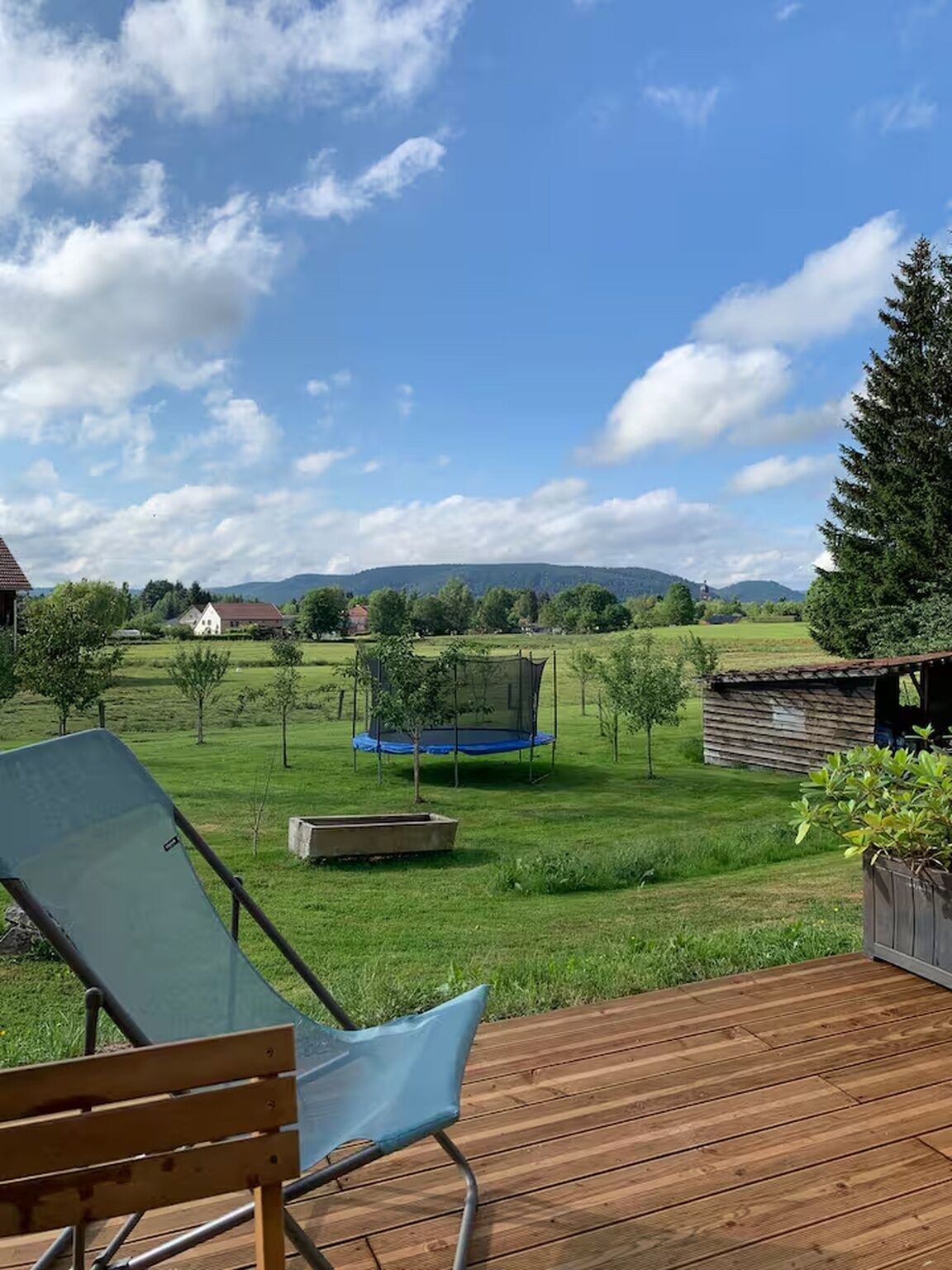 Une terrasse en bois avec une chaise en verre et un trampoline, le tout situé dans une cour verdoyante, avec des arbres et une colline en arrière-plan.