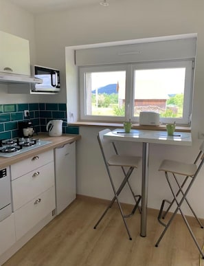 A compact kitchen with a microwave, kettle, and coffee maker on the counter, a window with a view of the outdoors, and a small dining table with two chairs.